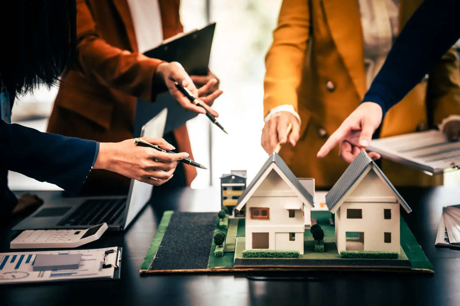 Asiatisches Immobilienteam in einer Diskussion mit zwei Männern und einer Frau, die sich auf ein Hausmodell auf einem Tisch konzentrieren, was auf eine Planungs- oder Verkaufssitzung hindeutet
